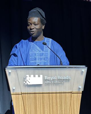 Dapo Bankole in a blue embroidered outfit and cap, speaking at a Royal Roads University podium.