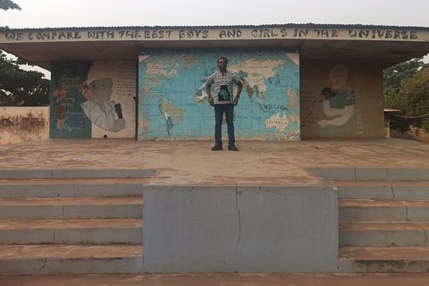 Dapo Bankole standing on the stage of the assembly ground at Mayflower School, Ikenne, under a painted sign reading We compare with the best boys and girls in the universe, between murals of the school's founders and a world map.
