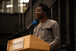 Dapo Bankole in a grey tunic and black cap, speaking at a Platform Calgary podium and looking off to the side.