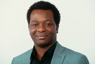 Landscape head and shoulders portrait of Dapo Bankole in a sage-green blazer and black shirt, smiling, against a light background.