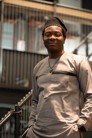 Dapo Bankole in a grey tunic and dark cap, smiling and leaning on a staircase railing.