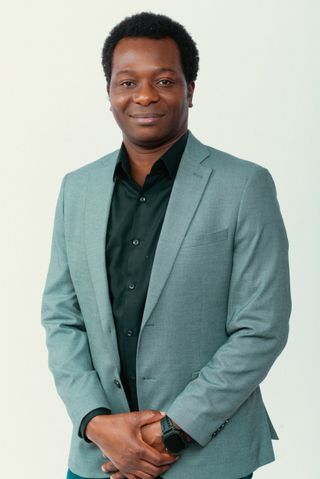 Dapo Bankole smiling, wearing a sage-green blazer over a black shirt, hands clasped, against a light studio background.