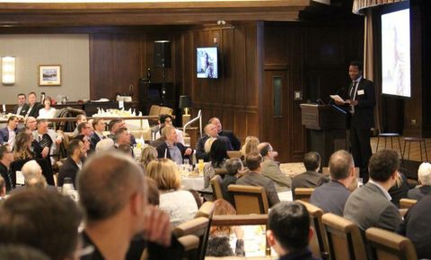Dapo Bankole speaking at a podium to a full room of guests seated at tables in a wood-panelled hall.