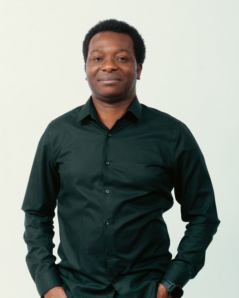 Dapo Bankole in a black shirt and teal jeans, hands in pockets, smiling against a light studio background.