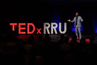 Dapo Bankole speaking on the TEDxRRU stage beside the large illuminated TEDxRRU letters, with the audience in silhouette.