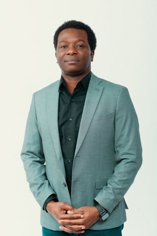 Dapo Bankole in a sage-green blazer and black shirt, hands clasped, with a calm expression, against a light studio background.