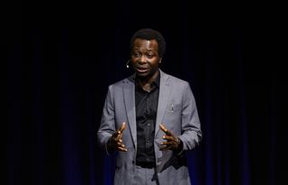Dapo Bankole mid-sentence on stage at TEDxRRU, wearing a grey suit and headset microphone, gesturing with both hands.