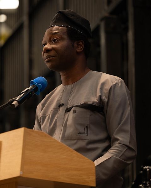 Dapo Bankole in a grey tunic and black cap, speaking at a Platform Calgary podium and looking off to the side.