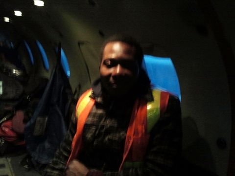Dapo Bankole in a high-visibility vest over a plaid shirt, inside the dim hold of a cargo plane, with cargo stacked behind him.
