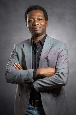Dapo Bankole in a grey blazer and black shirt with arms crossed, smiling, against a grey studio backdrop.